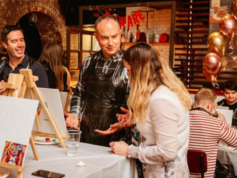 Three people conversing with paint on their hands, standing at a table with various art supplies